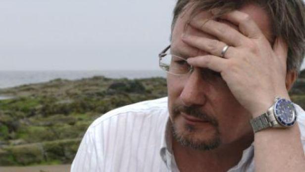 Ein Mann mit Brille und Uhr hält sich die Hand an die Stirn, im Hintergrund das Meer.