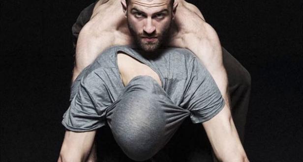 Ein muskulöser Mann hält eine andere Person fest, deren Kopf in einem grauen T-Shirt steckt.