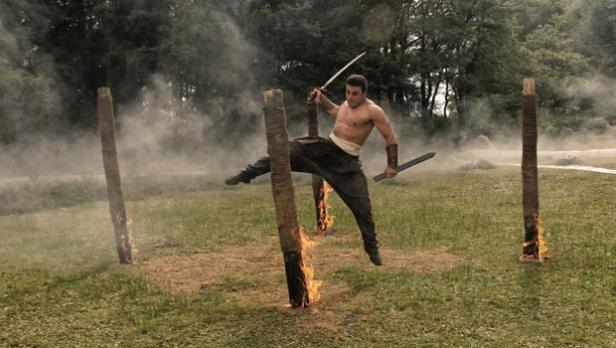 Ein Mann mit zwei Schwertern springt zwischen brennenden Holzpfosten hindurch.