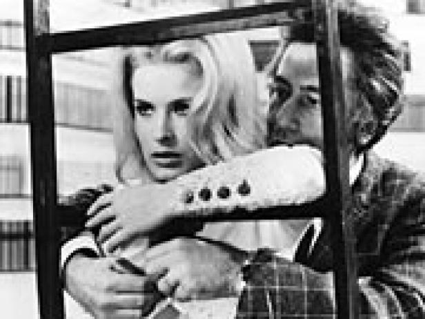 Eine blonde Frau wird von einem Mann umarmt, beide stehen hinter einem dunklen Rahmen.