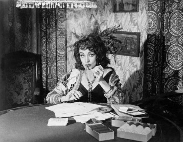 Eine Frau sitzt an einem Tisch mit Zigarren und Papieren, eine Zigarette im Mundwinkel.