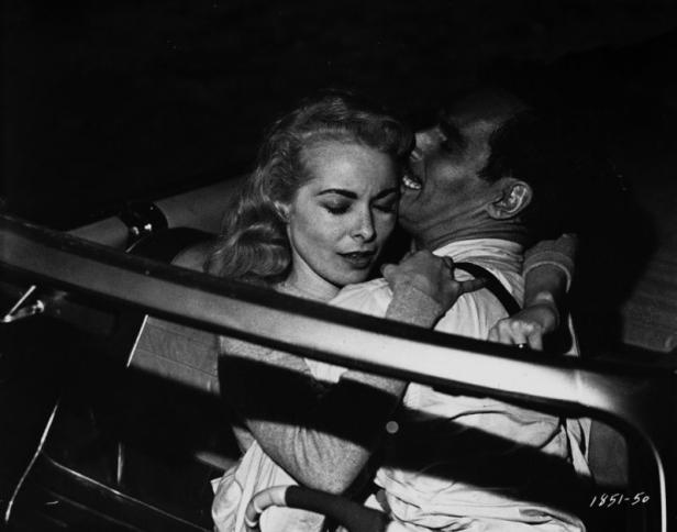 Ein Mann flüstert einer blonden Frau in einem Auto etwas ins Ohr.