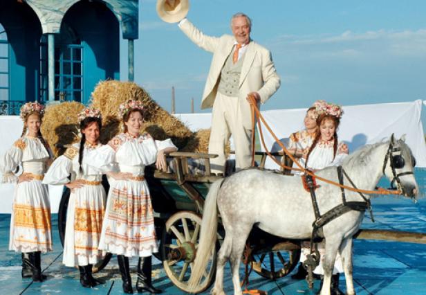Ein Mann im Anzug steht auf einem Heuwagen, der von einem Schimmel gezogen wird, umgeben von Frauen in Trachten.