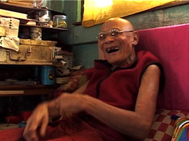 Ein älterer, glatzköpfiger Mann mit Brille lacht, während er auf einem Bett sitzt.