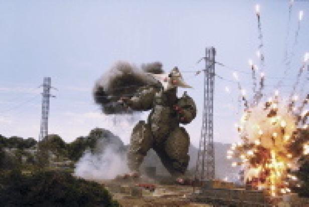 Ein riesiger Roboter steht inmitten von Explosionen und Strommasten.