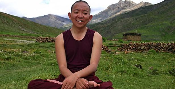 Ein lächelnder buddhistischer Mönch sitzt in einer Berglandschaft im Lotussitz.