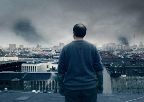 Ein Mann steht auf einem Dach und blickt auf die Skyline von Berlin mit Rauchwolken.