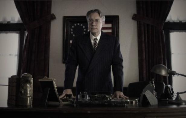 Ein Mann im Anzug steht an einem Schreibtisch vor einer amerikanischen Flagge.