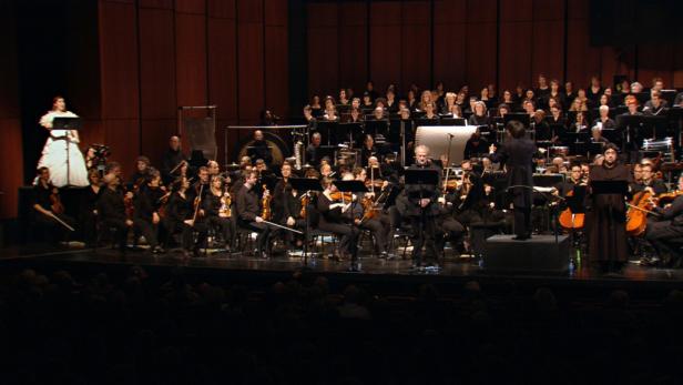 Ein Orchester und ein Chor treten auf einer Bühne auf, eine Sopranistin in Weiß steht links.