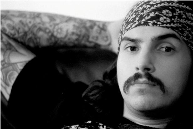 Ein Mann mit Tätowierungen, Schnurrbart und Kopftuch blickt in die Kamera.