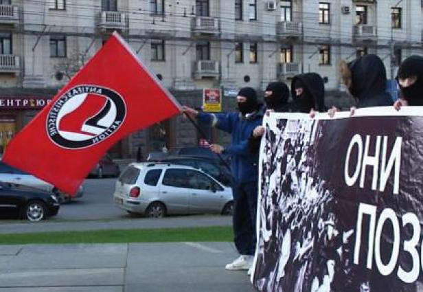 Vermummte Personen demonstrieren mit einer Antifa-Fahne und einem Banner.
