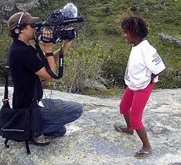Ein Kameramann filmt ein junges Mädchen in einem weißen T-Shirt und einer roten Hose.