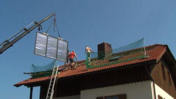 Ein Kran hebt ein Solarpaneel auf das Dach eines Hauses, wo Arbeiter stehen.