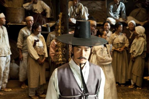 Ein Mann mit Hut und Schnurrbart steht vor einer Gruppe von Menschen in traditioneller koreanischer Kleidung.