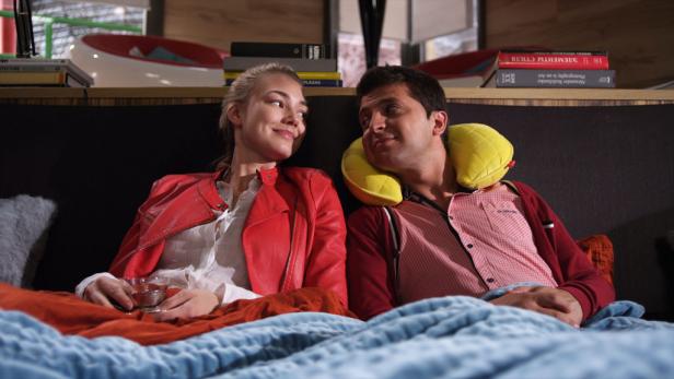 Ein Paar entspannt sich im Bett, die Frau hält ein Glas in der Hand, der Mann hat ein Nackenkissen.