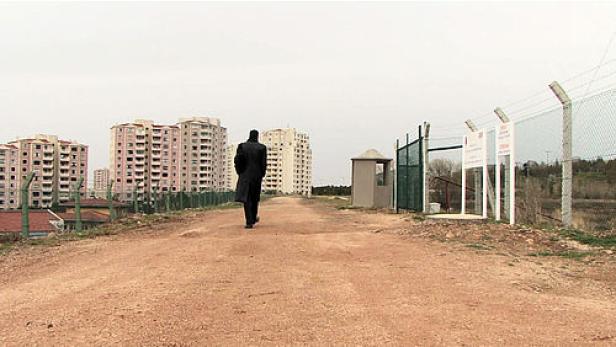 Ein Mann in einem Mantel geht auf einer unbefestigten Straße in Richtung einer Wohnsiedlung.