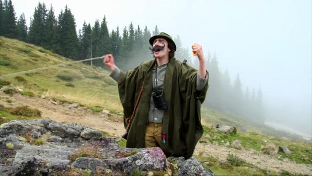 Ein Mann mit Hut und falschem Schnurrbart steht jubelnd in den Bergen.