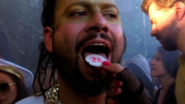 Ein Mann mit Make-up hält eine Pille mit der Zahl 25 auf seiner Zunge.