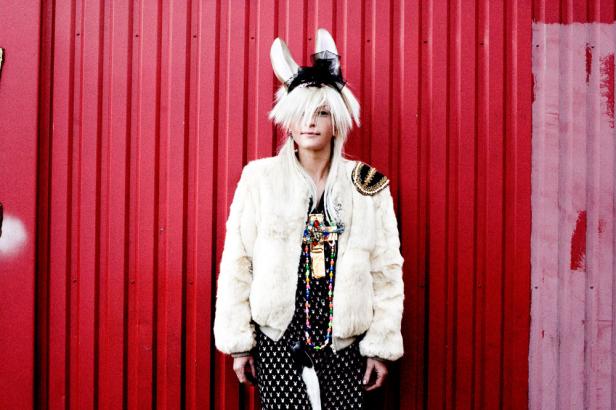 Eine Person mit Hasenohren, Pelzjacke und auffälligem Schmuck vor einer roten Wand.