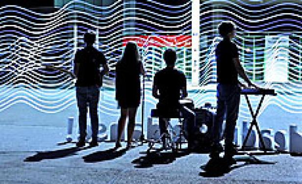 Eine Band spielt im Freien vor einem Gebäude mit wellenförmigen Lichtprojektionen.