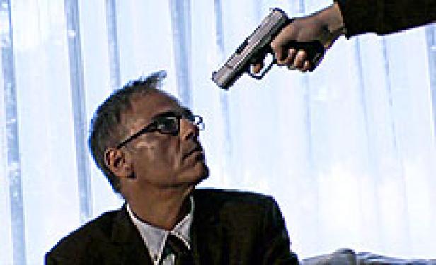 Ein Mann mit Brille wird mit einer Pistole bedroht.