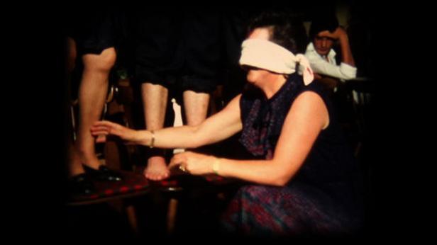 Eine Frau mit verbundenen Augen ertastet die Beine von Personen, die auf Stühlen sitzen.