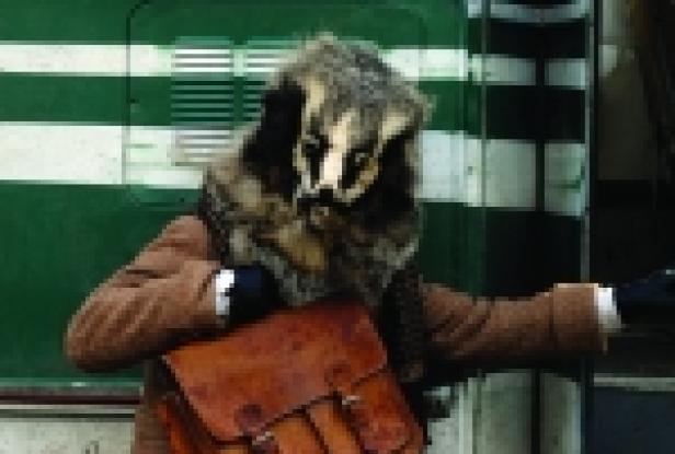 Eine Person in einem Dachskostüm steht vor einem grünen Zug und trägt eine braune Tasche.