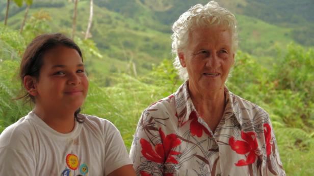 Ein Mädchen und eine ältere Frau sitzen lächelnd vor einer grünen Landschaft.