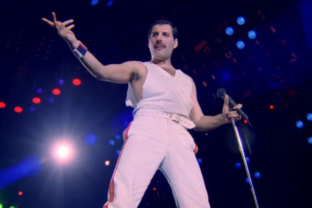 Freddie Mercury von Queen tritt in weißer Kleidung mit einem Mikrofon in der Hand auf einer Bühne auf.