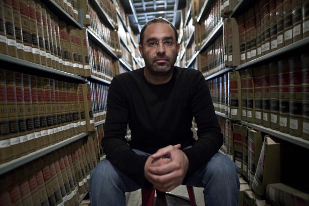 Ein Mann mit Brille sitzt in einem Raum voller Bücherregale.