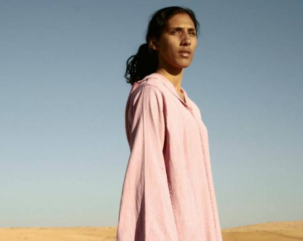 Eine Frau in einem rosa Gewand steht in der Wüste und blickt nach oben.