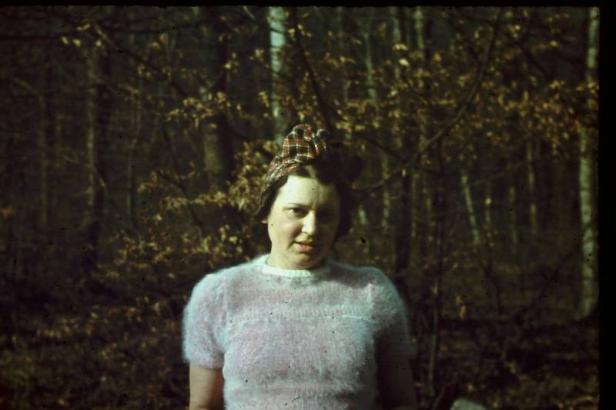 Eine Frau mit kariertem Hut und rosa Pullover steht vor einem Wald.