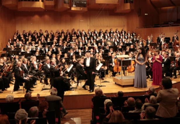 Ein Orchester und ein Chor treten auf einer Bühne vor Publikum auf.