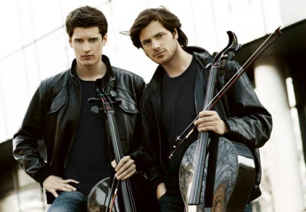 Zwei Männer in Lederjacken posieren mit ihren Celli vor einem Gebäude.