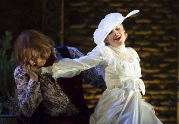 Ein Mann küsst die Hand einer Frau in einem weißen Kleid und Hut während einer Bühnenaufführung.