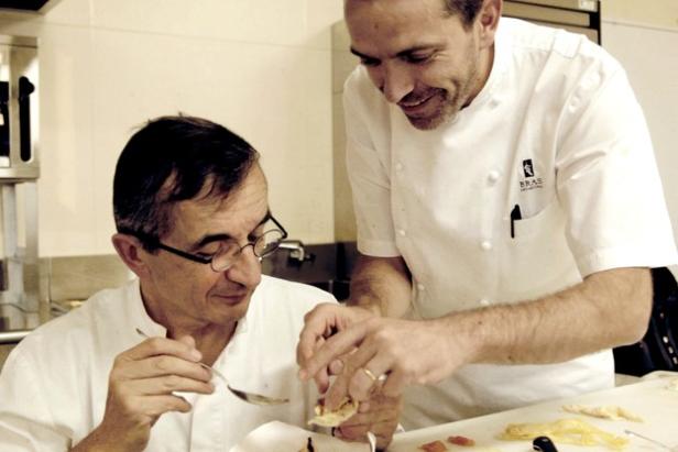 Zwei Köche bereiten in einer Küche Speisen zu, einer probiert mit einem Löffel.