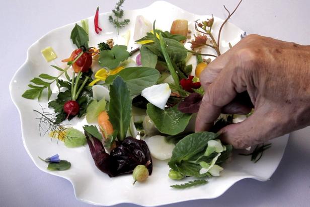 Eine Hand drapiert essbare Blüten und Kräuter auf einem weißen Teller.
