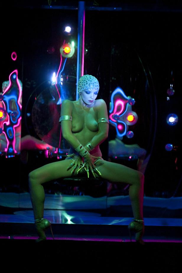 Eine Frau mit silbernen Armreifen und Kopfbedeckung posiert auf einer Bühne mit Neonlicht.