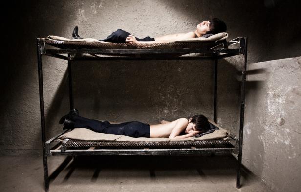 Zwei Männer liegen in einer kargen Zelle auf einer Doppelstockliege.