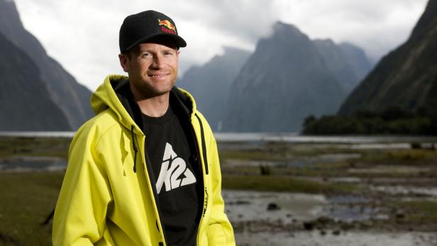 Ein Mann mit Kappe und gelber Jacke vor einer Berglandschaft.