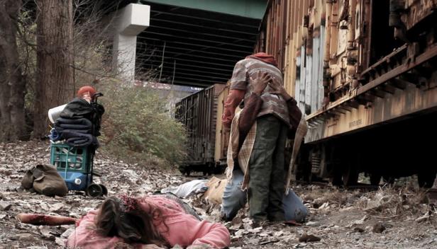 Eine leblose Frau liegt auf Bahngleisen, während eine Person von hinten umarmt wird.