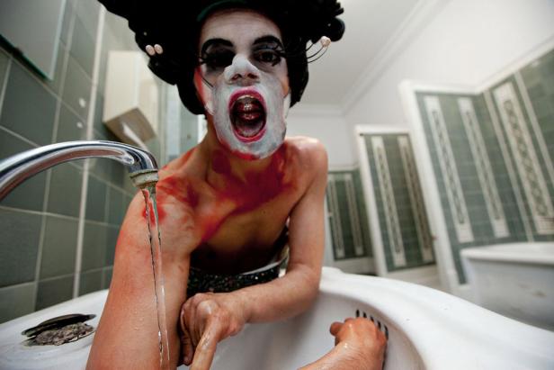 Eine Person mit Make-up und theatralischer Kleidung steht an einem Waschbecken mit laufendem Wasser.
