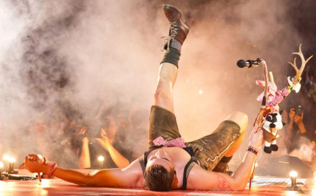 Ein Mann in Lederhosen liegt auf der Bühne und hält ein dekoriertes Mikrofonstativ in der Hand.
