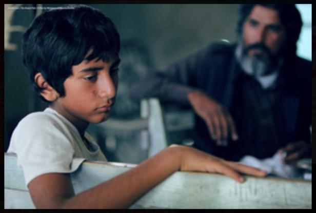 Ein nachdenklicher Junge sitzt an einem Tisch, im Hintergrund ein Mann mit Bart.