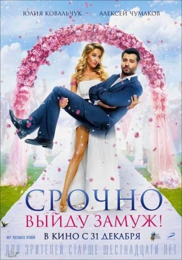 Ein Mann trägt eine Frau im Brautkleid vor einem Rosenbogen.