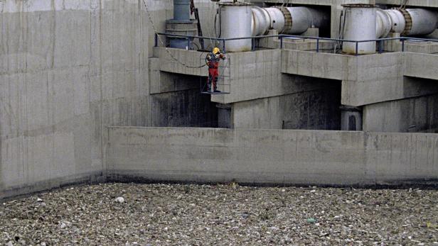 Ein Arbeiter in Schutzkleidung steht auf einer Plattform vor einer Betonwand und Industrieanlagen.