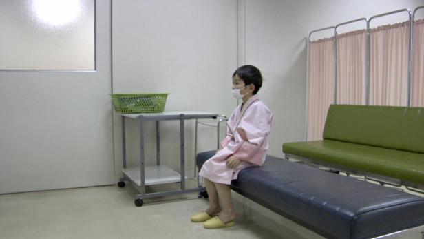 Ein Kind mit Maske und rosa Morgenmantel sitzt in einem Wartezimmer.