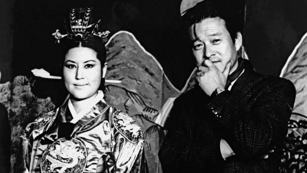 Eine Frau in traditioneller koreanischer Kleidung und ein Mann im Anzug posieren vor einem bemalten Hintergrund.