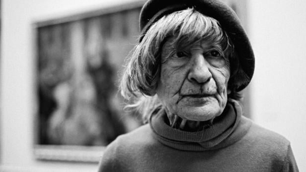 Schwarzweißporträt einer älteren Frau mit Hut vor einem unscharfen Gemälde.