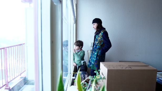 Eine Frau und ein Kind schauen aus dem Fenster eines Zimmers mit Umzugskartons.